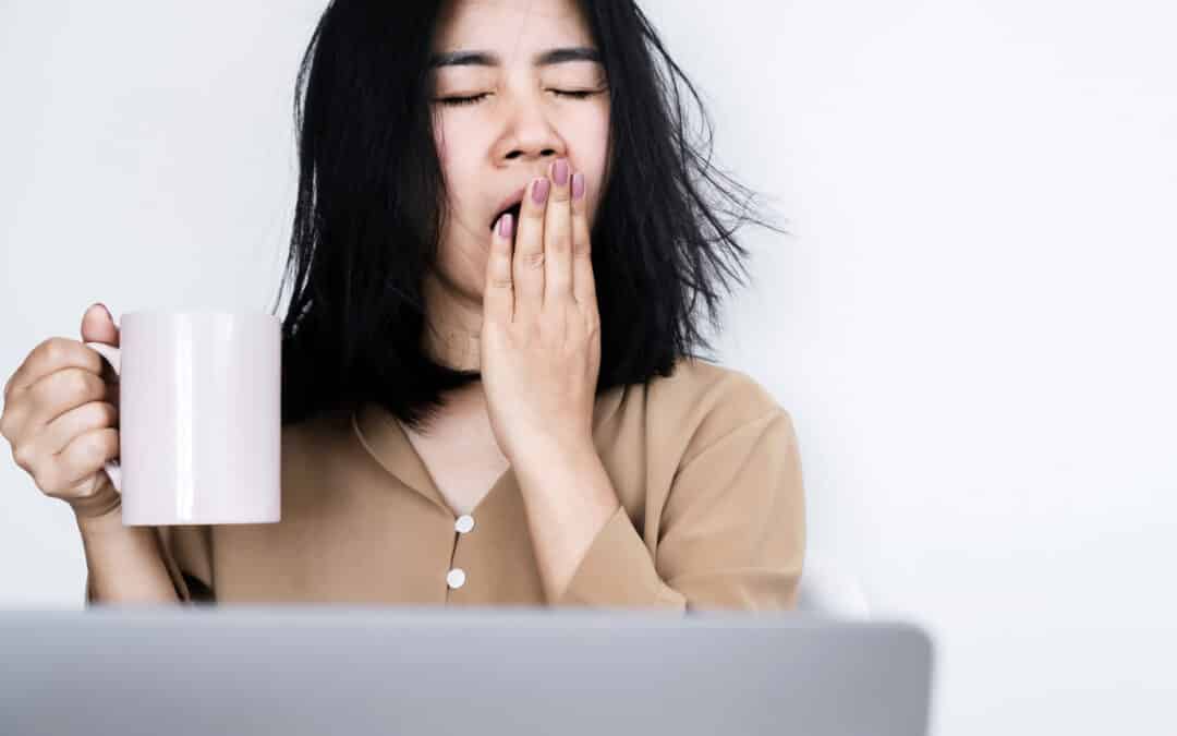 overworked Asian businesswoman yawning at office desk hand holding a cup of coffee feeling tired and sleepy