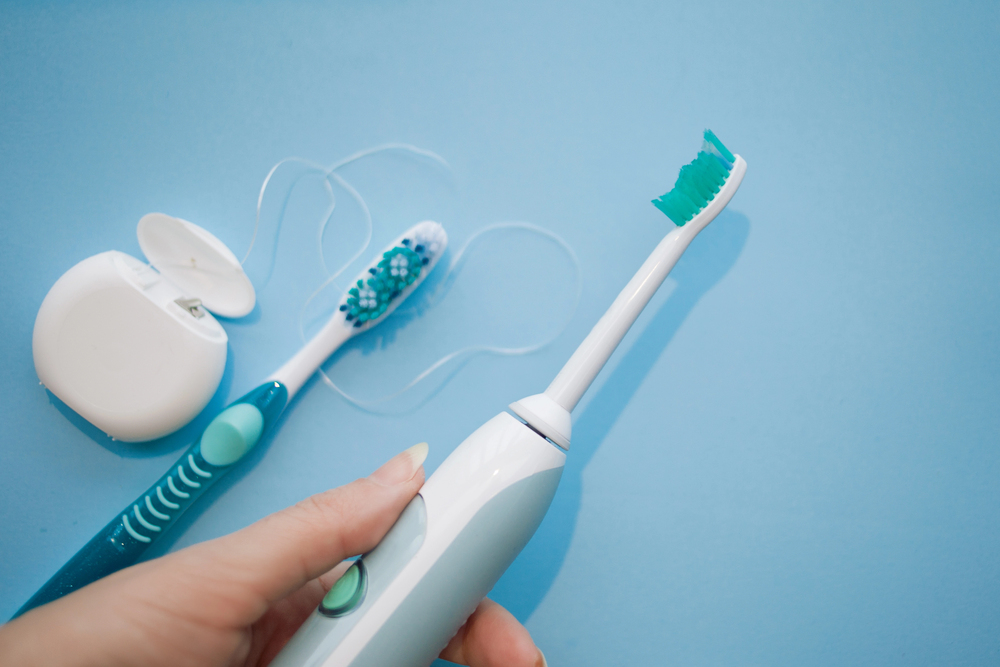 woman holding an electric toothbrush with a manual toothbrush and floss sitting on table beneath
